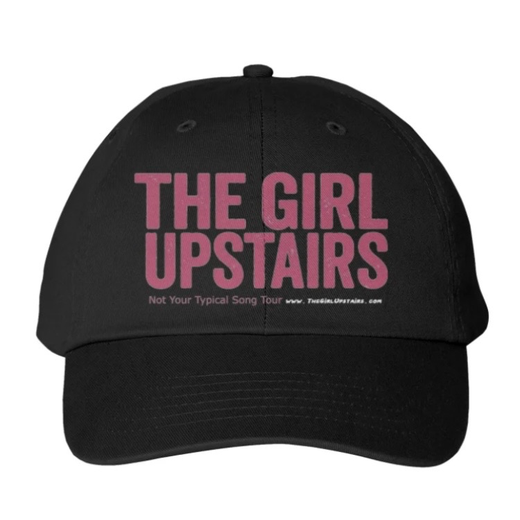 Black baseball cap with pink “THE GIRL UPSTAIRS” text and small slogan beneath.