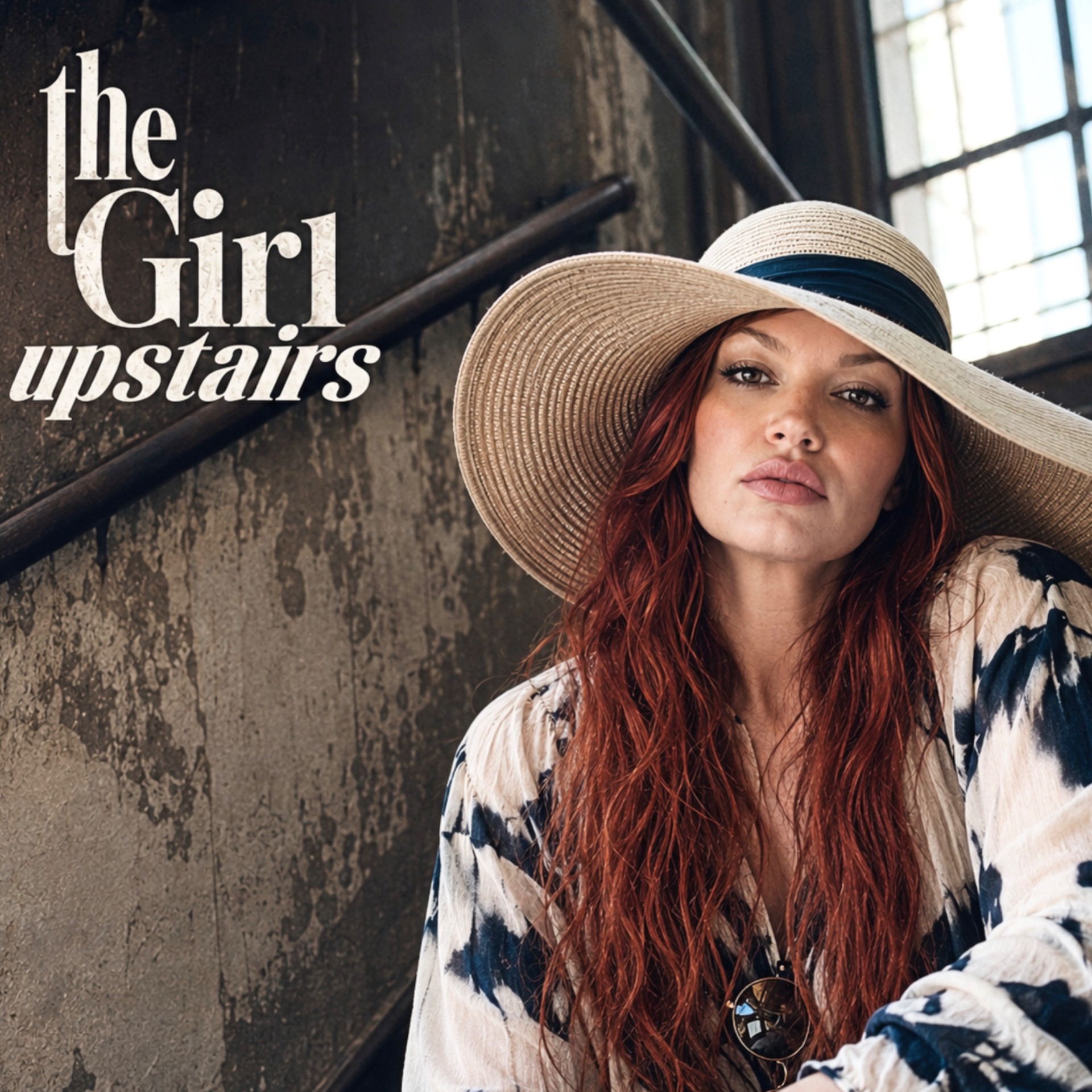 music artist, The Girl Upstairs wearing a wide straw hat and patterned shirt, posing on a staircase in an industrial building